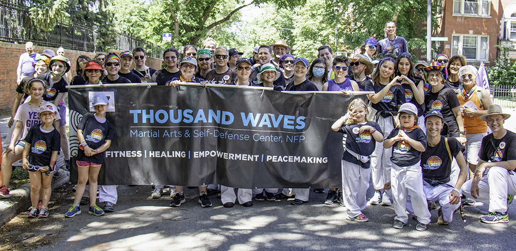 Members of Thousand Waves behind a banner with the text Thousand Waves Martial Arts & Self-Defense Center, NFP Fitness | Healing | Empowerment | Peacemaking